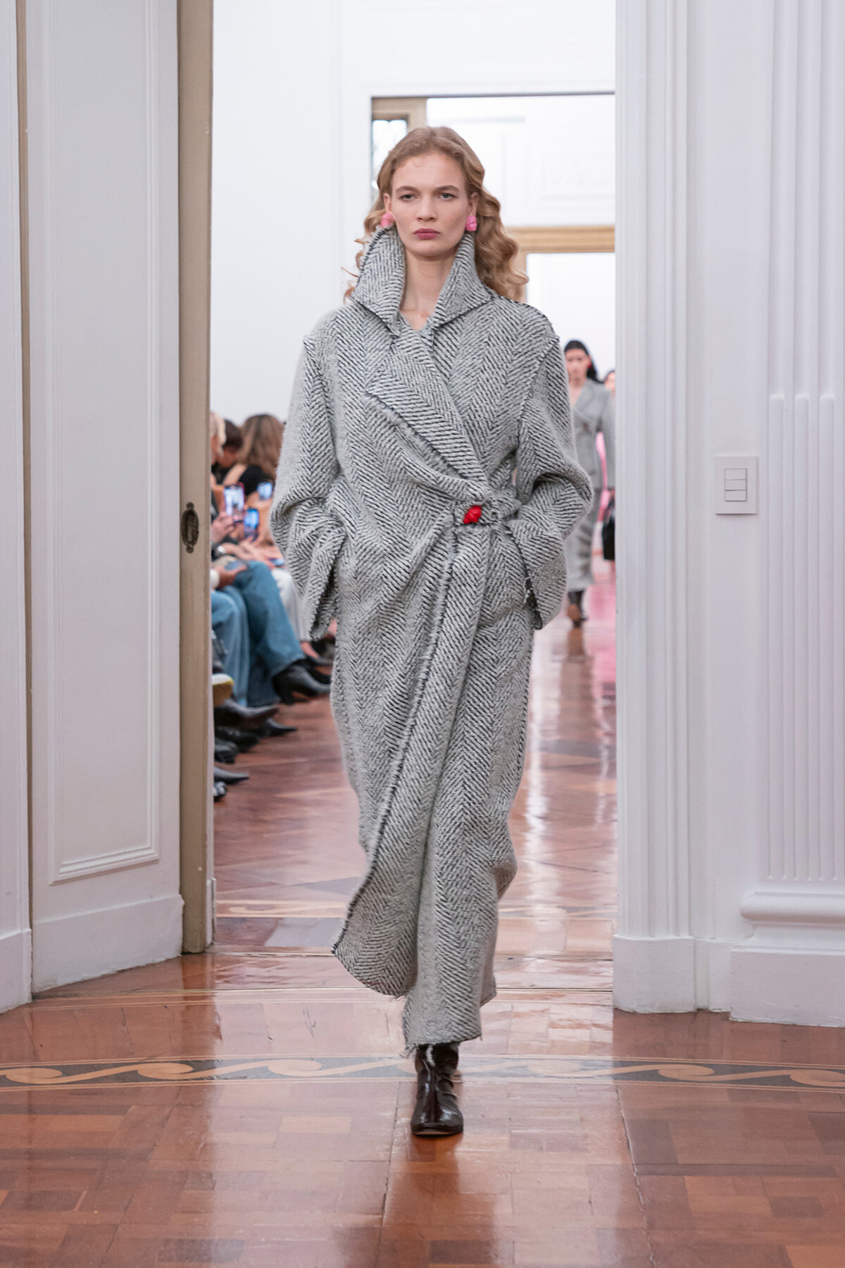 Model in a gray herringbone wrap coat walking a runway with hands in pockets and a red waist accent, black boots visible at the end of the coat. Audience watches from both sides in a bright room.
