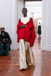 Model walks a fashion show wearing a bright red cape draped over a white sleeveless top and ivory satin wide-leg pants.
