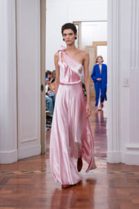 Model in a pink satin one-shoulder gown walks the runway in a bright, classical room; audience seated along the sides.