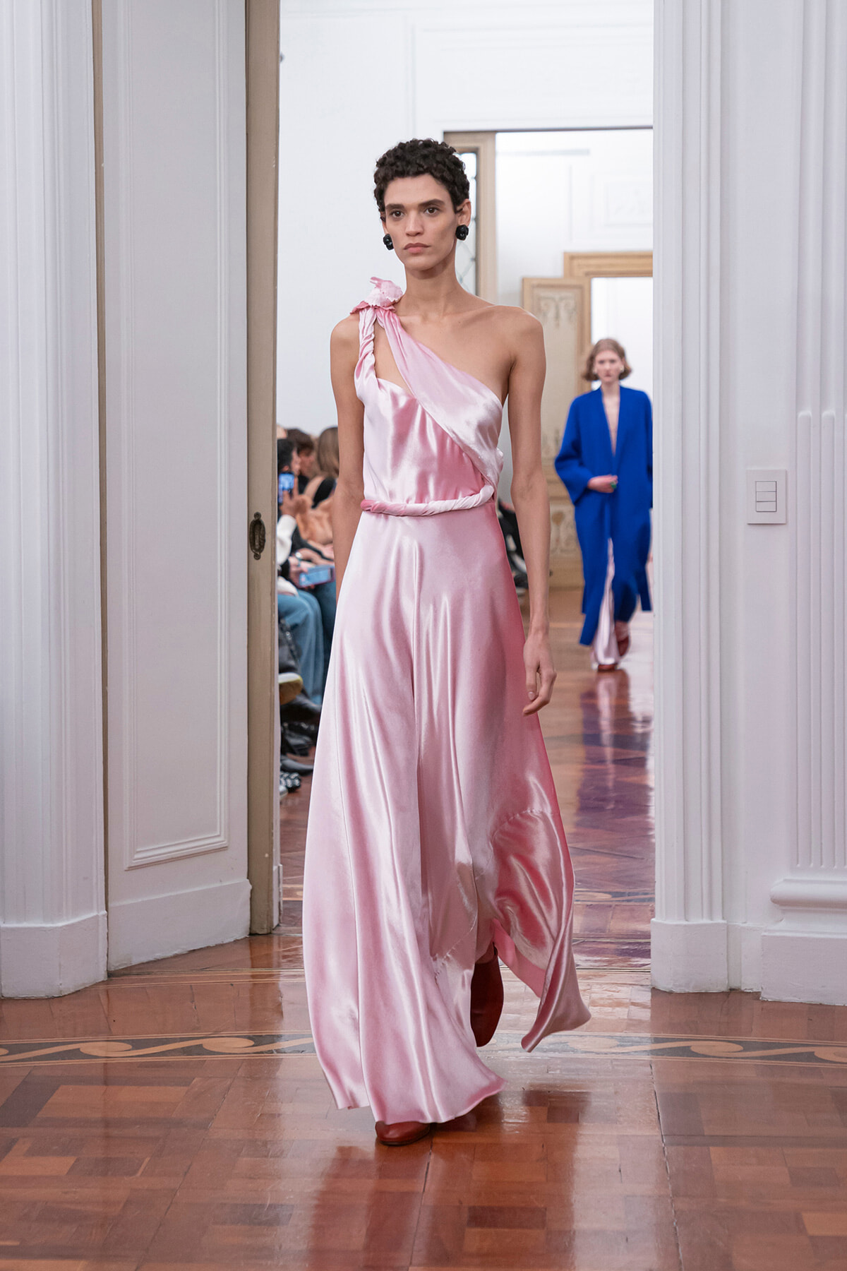Model in a pink satin one-shoulder gown walks the runway in a bright, classical room; audience seated along the sides.