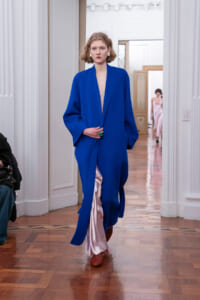 Model in a vivid blue oversized coat with pink satin lining peeking at the front, walking a runway in a wood-floored room.