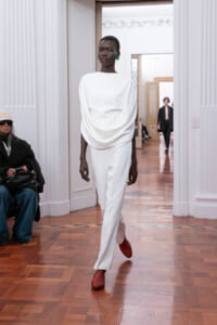 Model in an all-white, draped top and white pants walks the runway in a bright hall, wearing green earrings and red shoes.