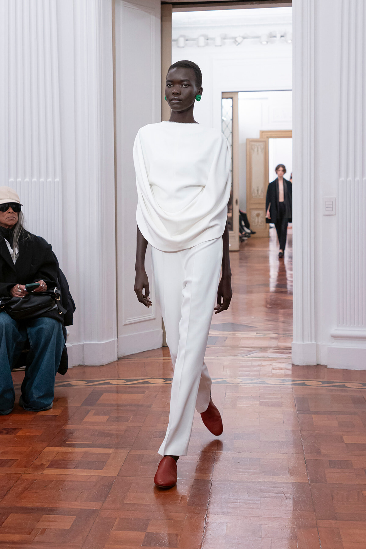Model in an all-white, draped top and white pants walks the runway in a bright hall, wearing green earrings and red shoes.