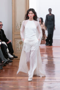 Model walks a runway in an all-white outfit with a sheer-sleeve top and wide-leg pants, red statement earrings, in a wooden-floored venue.