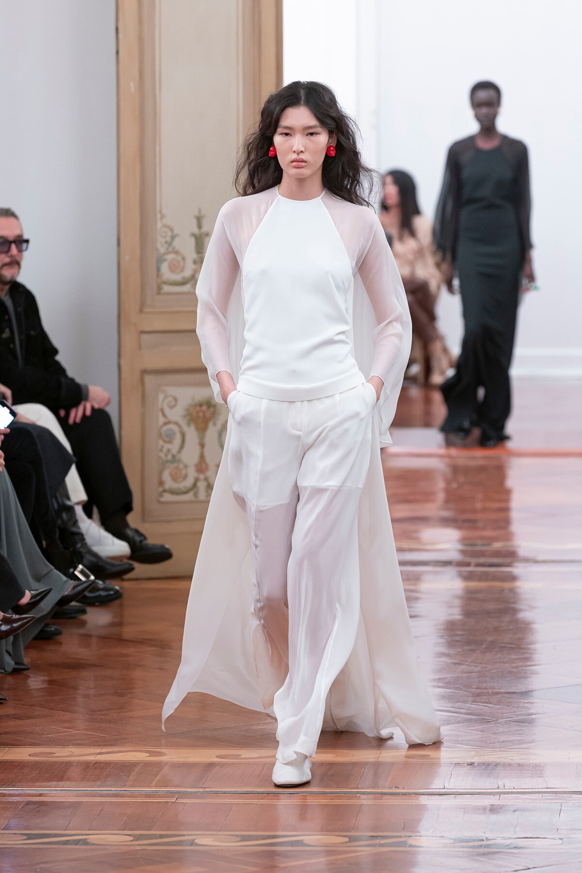 Model walks a runway in an all-white outfit with a sheer-sleeve top and wide-leg pants, red statement earrings, in a wooden-floored venue.