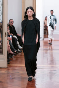 Model walks the runway in a black outfit with a beaded cape, hands in pockets, wearing black loafers; audience sits along the left.