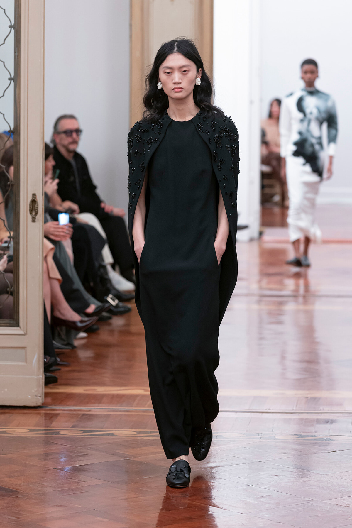 Model walks the runway in a black outfit with a beaded cape, hands in pockets, wearing black loafers; audience sits along the left.