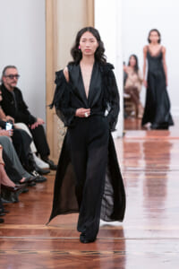 Model walks a fashion show in an all-black outfit with sheer wide-leg pants and ruffled cape sleeves, audience in the background.