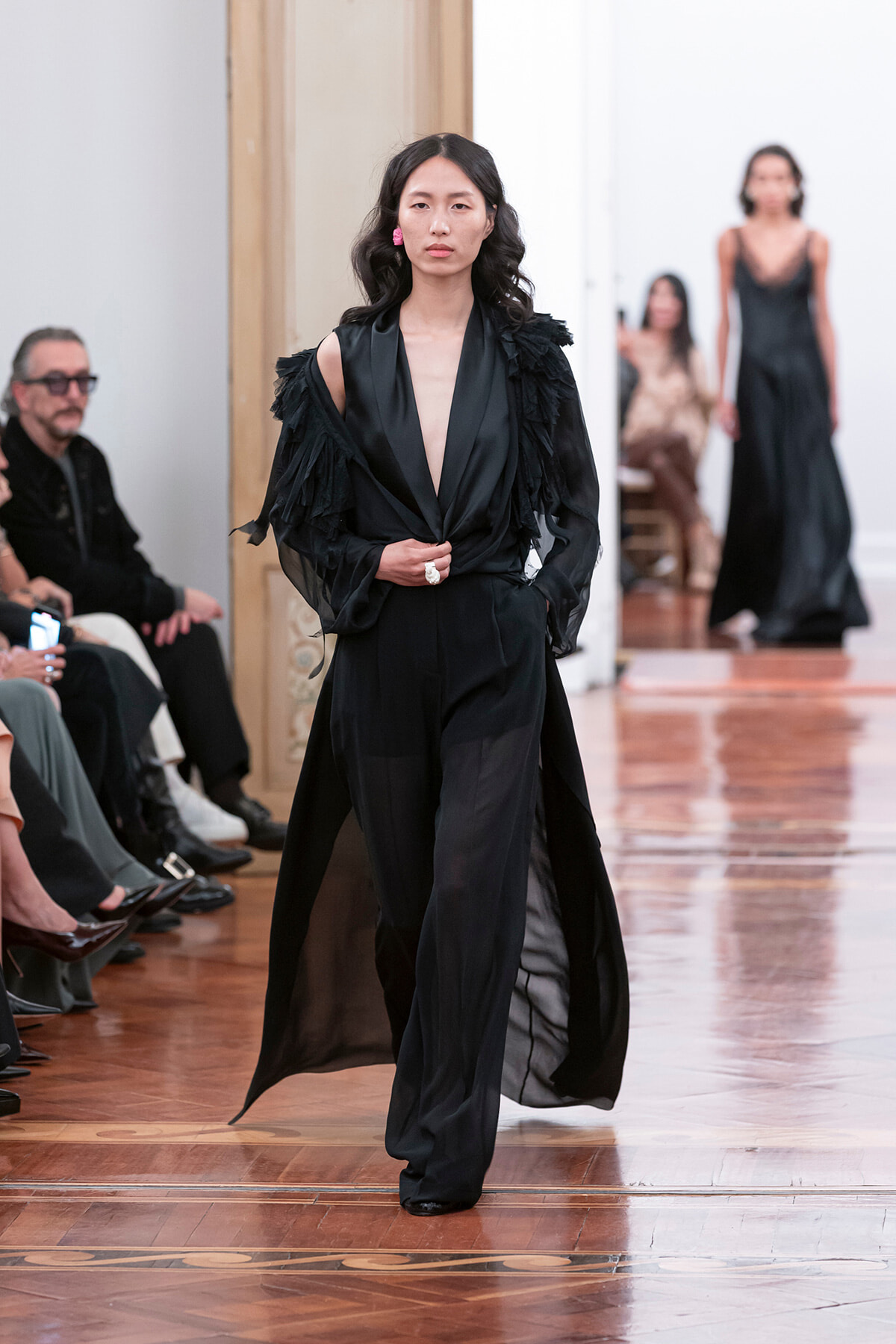 Model walks a fashion show in an all-black outfit with sheer wide-leg pants and ruffled cape sleeves, audience in the background.