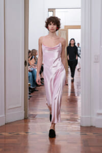 Model walks a pale pink satin slip dress down a runway with seated audience in a bright room behind her.
