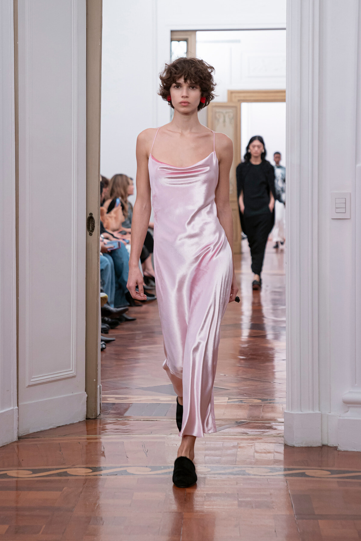Model walks a pale pink satin slip dress down a runway with seated audience in a bright room behind her.