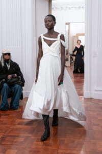 Model walks a white draped gown with twisted strap bodice and knee-high black boots on a wooden runway.