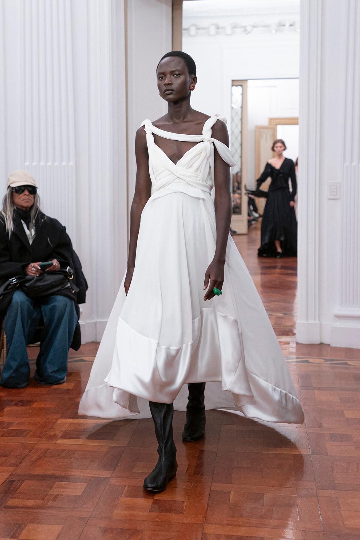Model walks a white draped gown with twisted strap bodice and knee-high black boots on a wooden runway.