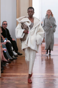 Runway scene: model wearing an oversized white fur coat over light gray top and cream trousers, carrying a taupe bag.