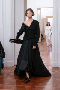 Model walking in a black, one-shoulder gown with floral bodice and a large black clutch.