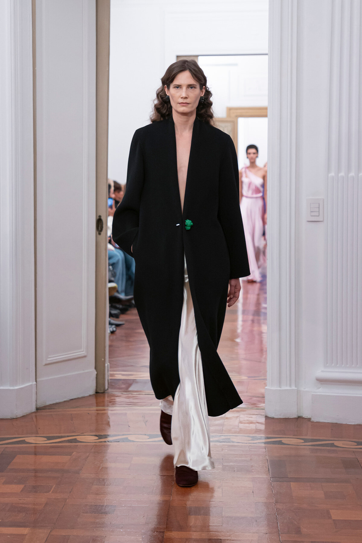 Model walks down a runway in a black long coat with a deep V opening, white satin pants peeking below, and brown shoes; audience seen in a bright room behind.