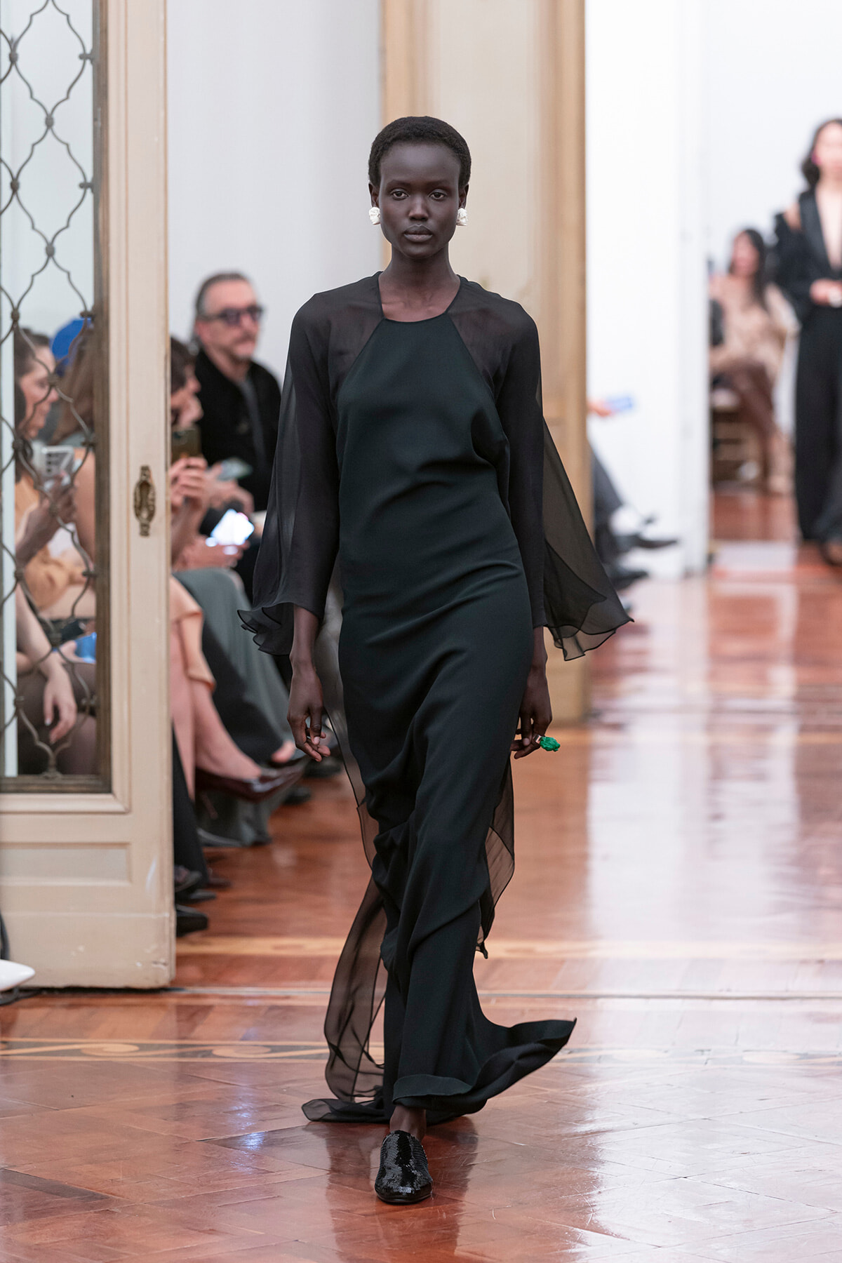 Model walks a fashion runway wearing a black floor-length gown with sheer, flowing sleeves and statement earrings.
