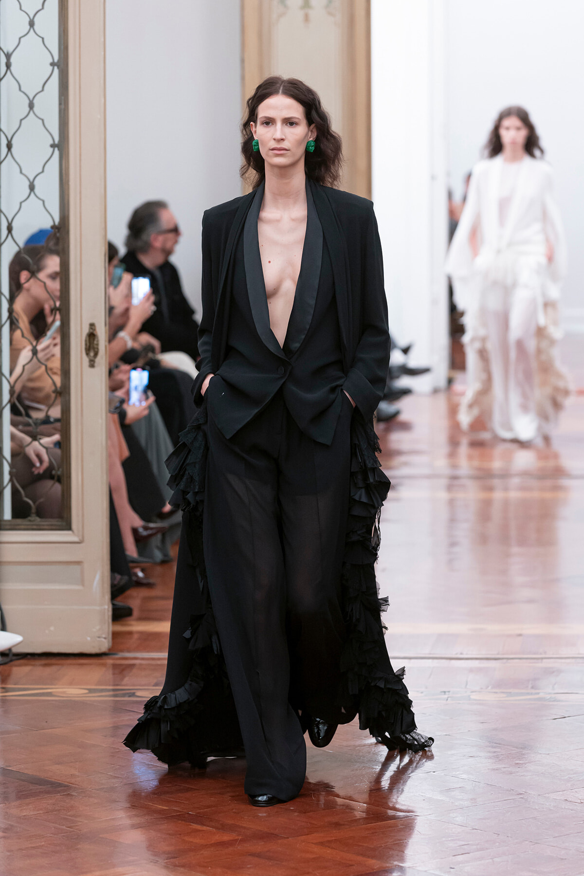Model walking a runway in a black deep-plunge blazer and wide-leg pants with ruffled sides; audience watches from the left.