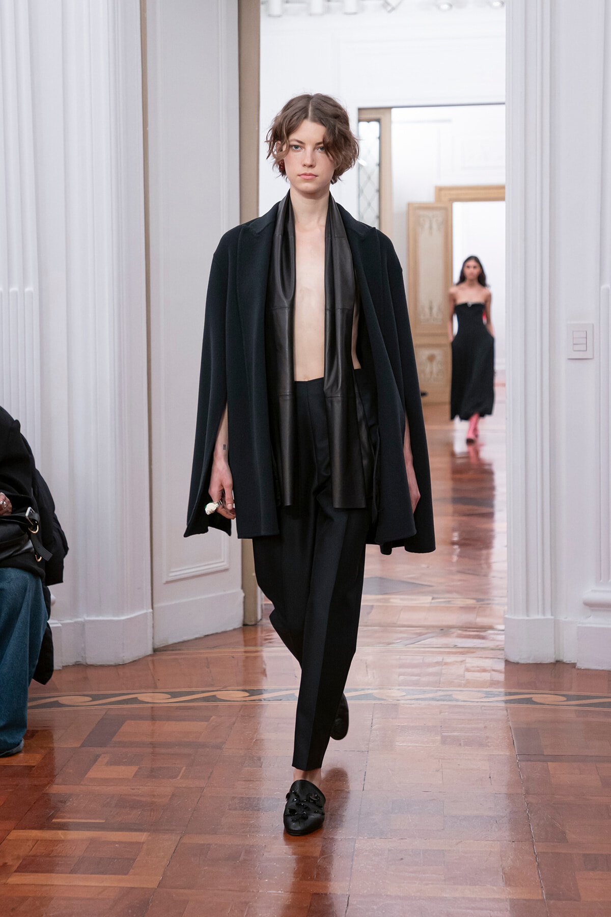 Model walking a fashion show in an all-black outfit: loose cape over a black leather scarf, cropped top, tailored pants, and black loafers.