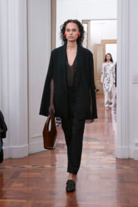 Model in a black suit with a sheer top, red earrings, and a brown handbag, walking down a wooden-floored hallway runway.