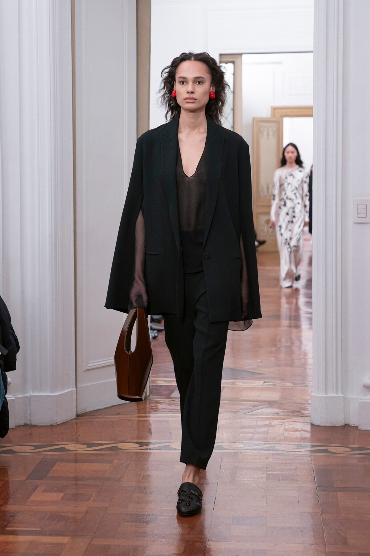 Model in a black suit with a sheer top, red earrings, and a brown handbag, walking down a wooden-floored hallway runway.