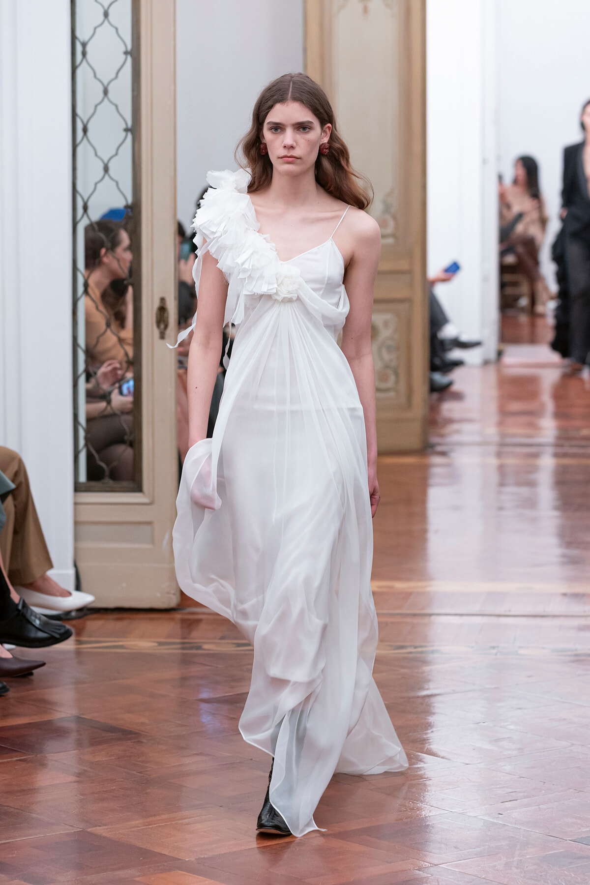 Model walks a runway in a white one-shoulder chiffon gown with a large ruffle, indoors on a wood floor show set.