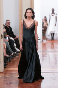 Model walks the runway in a black satin slip dress with lace V-neck; seated audience along the catwalk.