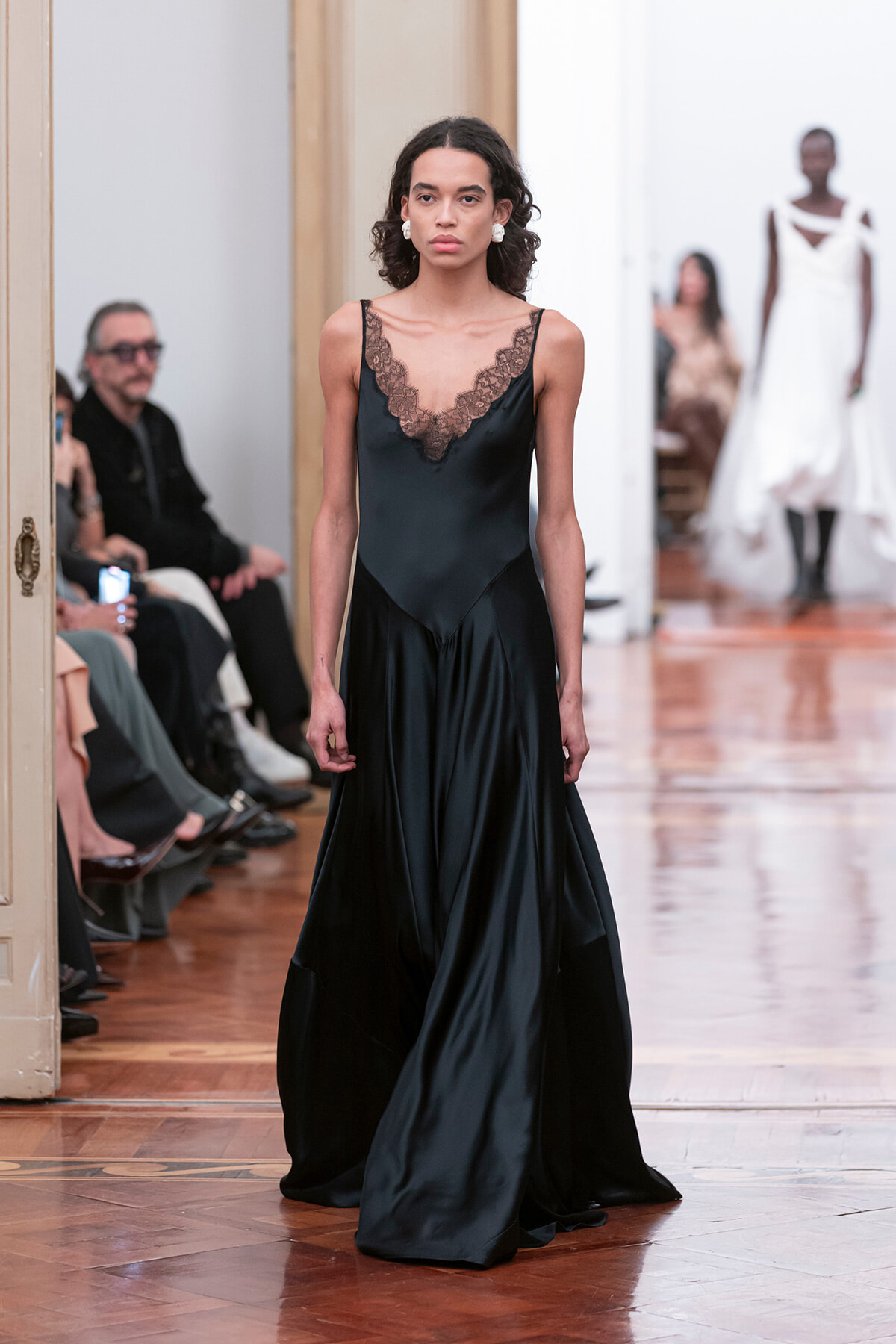 Model walks the runway in a black satin slip dress with lace V-neck; seated audience along the catwalk.