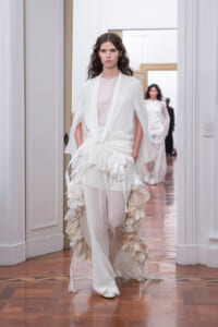 Model walking a white, layered gown with ruffled cascading accents at a fashion show.