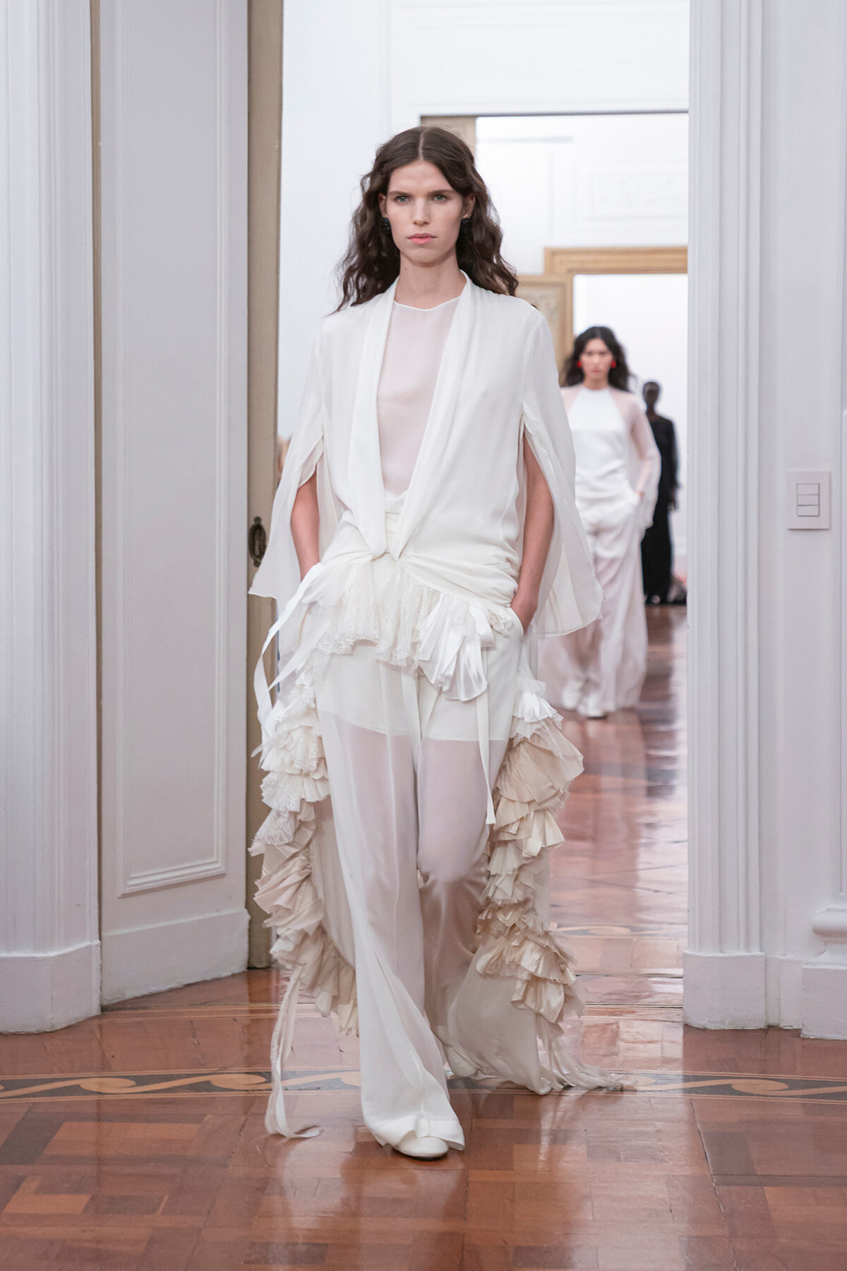 Model walking a white, layered gown with ruffled cascading accents at a fashion show.