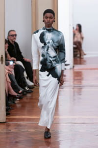 Model walks a white maxi dress with a bold black abstract print at a fashion show runway, high neckline and long sleeves.