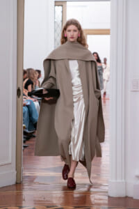 Model walking a fashion show in a taupe cape coat over a white satin dress, holding a black clutch.