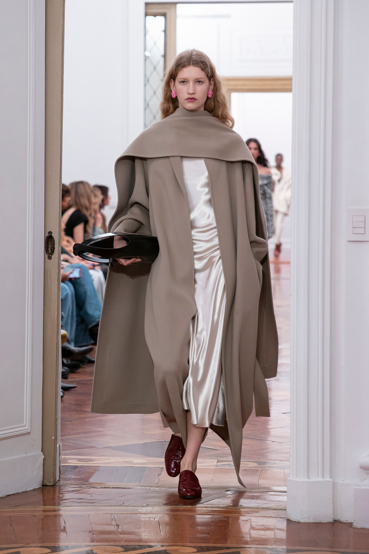 Model walking a fashion show in a taupe cape coat over a white satin dress, holding a black clutch.