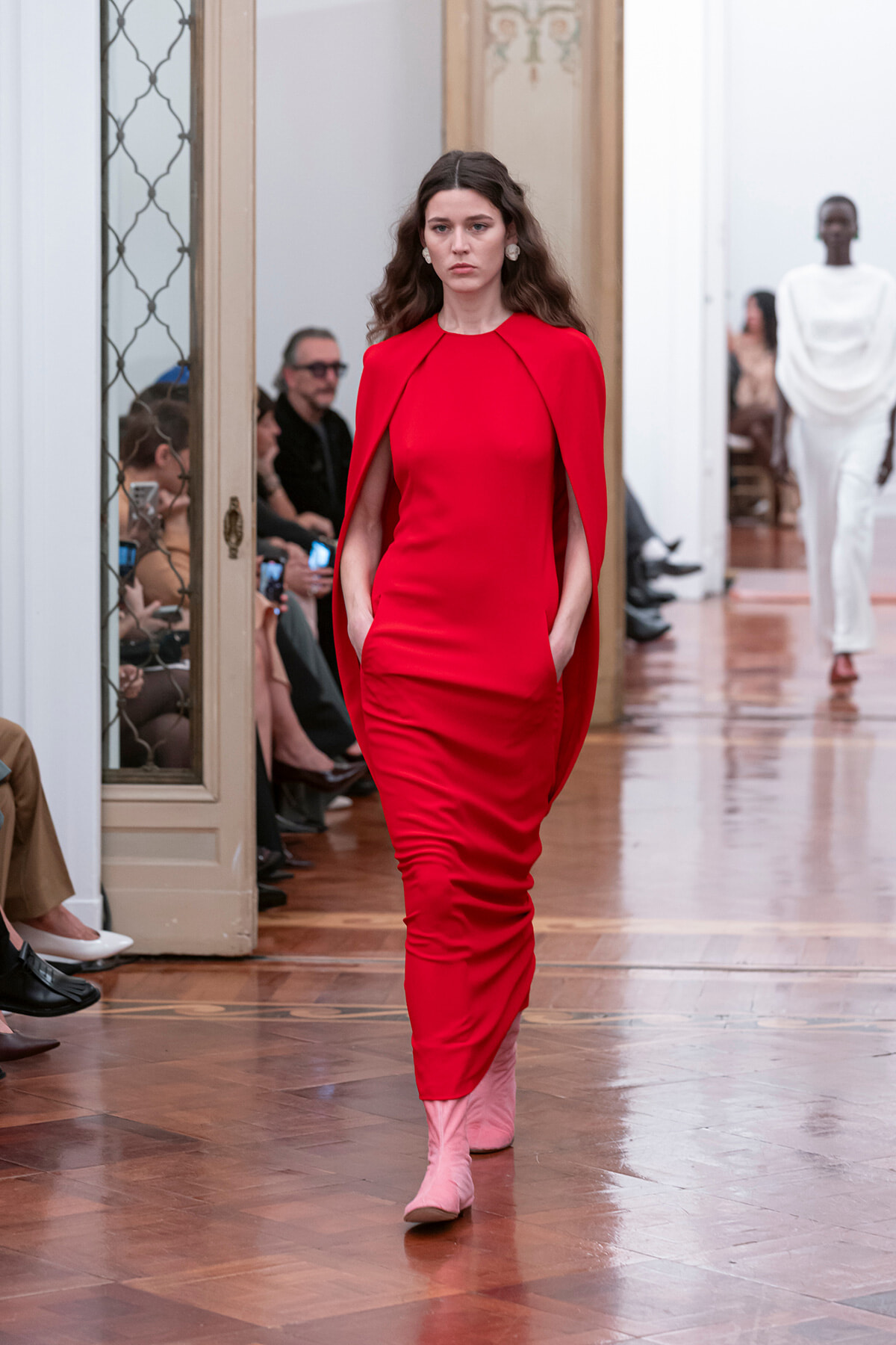 Model walks the runway in a bold red cape‑dress with pockets, paired with pink ankle boots.