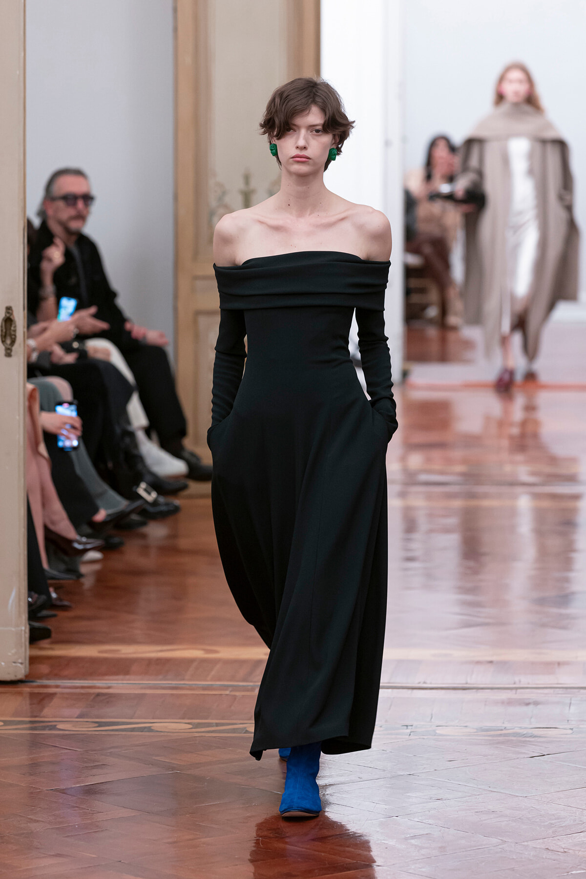 Model on a fashion runway wearing an off-the-shoulder black gown with blue ankle boots and green earrings.