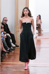 Female model walks a runway in a strapless black jumpsuit with silver trim at the top, hands in pockets, wearing pink socks or boots.