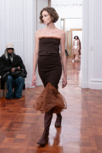 Model walks a runway in a strapless dark brown dress with a voluminous sheer brown hem and knee-high boots; audience sits along the wall in a gallery setting.
