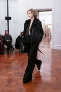 Model striding down a runway in a black ruffled cape and wide-leg pants, green earrings, in a white-paneled venue with wooden floor.
