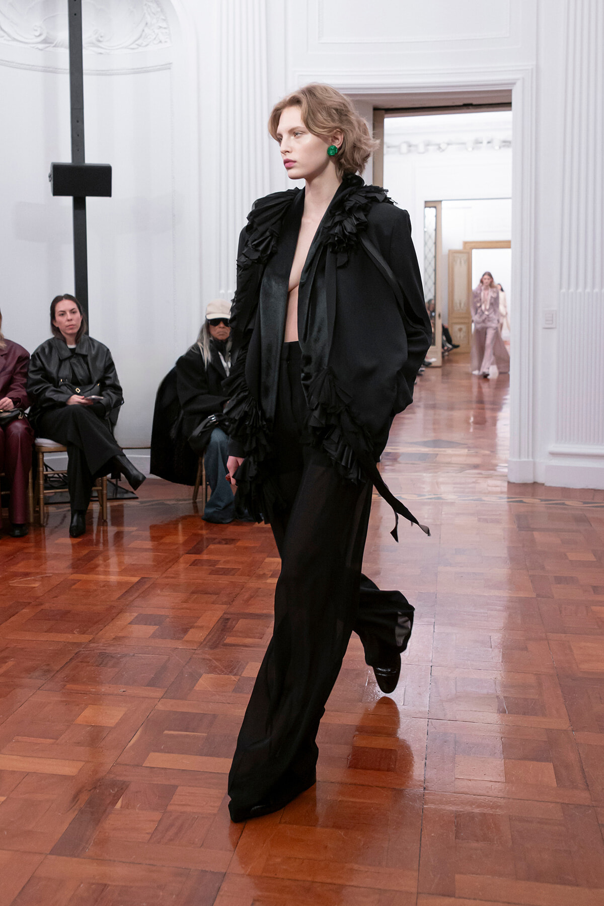 Model striding down a runway in a black ruffled cape and wide-leg pants, green earrings, in a white-paneled venue with wooden floor.
