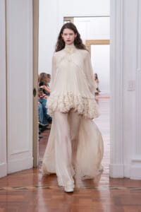 Model walking a runway in an ivory, high-neck gown with cascading ruffles and sheer sleeves.