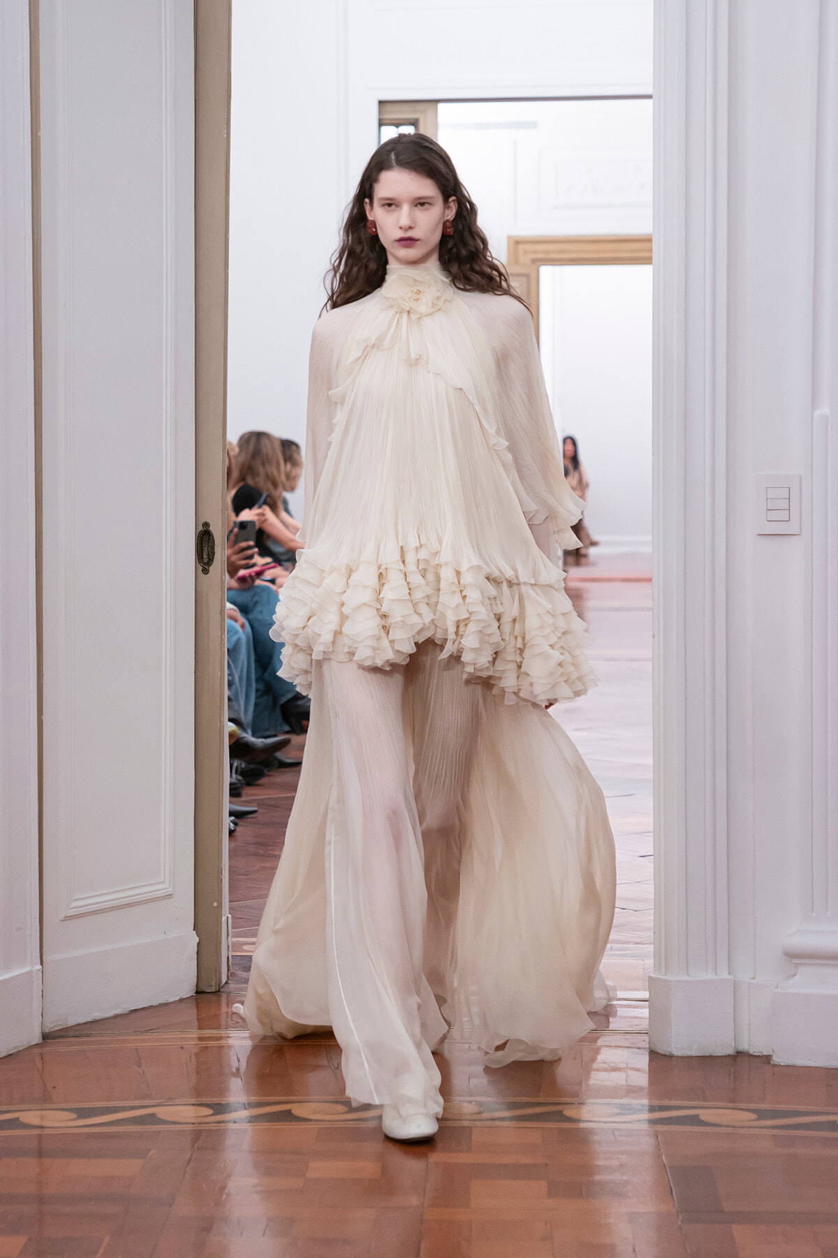 Model walking a runway in an ivory, high-neck gown with cascading ruffles and sheer sleeves.