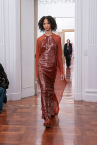 Model walks a runway in a rust-colored sequin gown with a sheer red cape in a bright gallery setting.