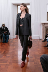 Model on a runway wearing a dark, tailored suit with a beaded crop top and red loafers, holding a small dark bag in a white-walled room.