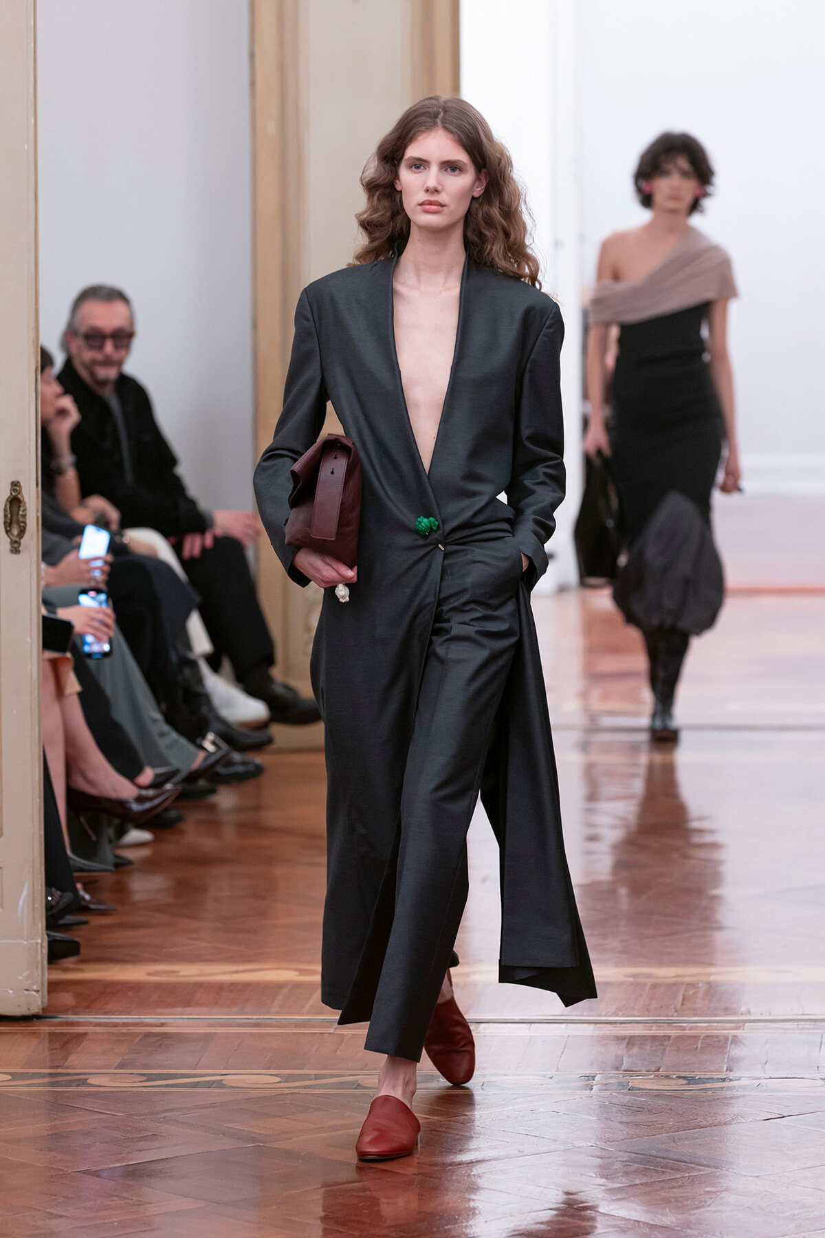 Model walks a runway in a dark plunging blazer and matching wide-leg pants, carrying a burgundy clutch and wearing red loafers.
