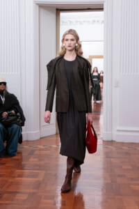 Model walking a runway in a black blazer over a black dress, brown knee-high boots, and a red handbag on a wooden-floor venue.