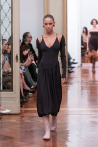 Model walking the runway in a black midi dress with cutouts at the shoulders and long sleeves, paired with pale pink boots, in a gallery-style venue.