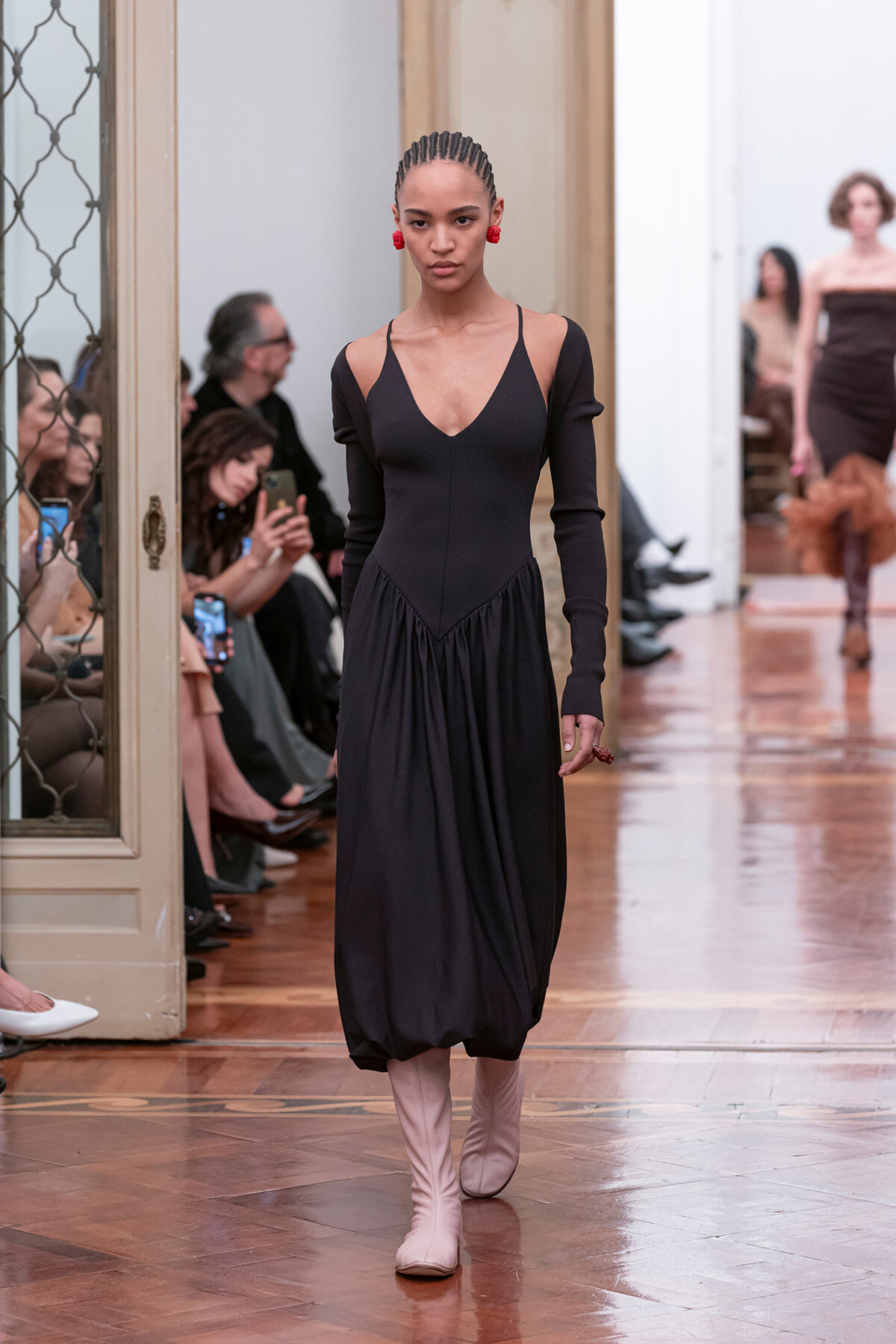 Model walking the runway in a black midi dress with cutouts at the shoulders and long sleeves, paired with pale pink boots, in a gallery-style venue.