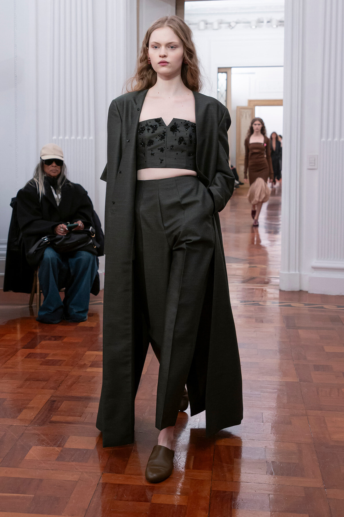 Model walks a runway in a dark, oversized coat over a beaded black crop top and wide-leg trousers on a polished wood floor.