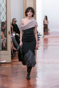 Model walks the runway in a dark, fitted dress with a pale pink off-shoulder drape and a sculpted gray bubble hem, carrying a black handbag.