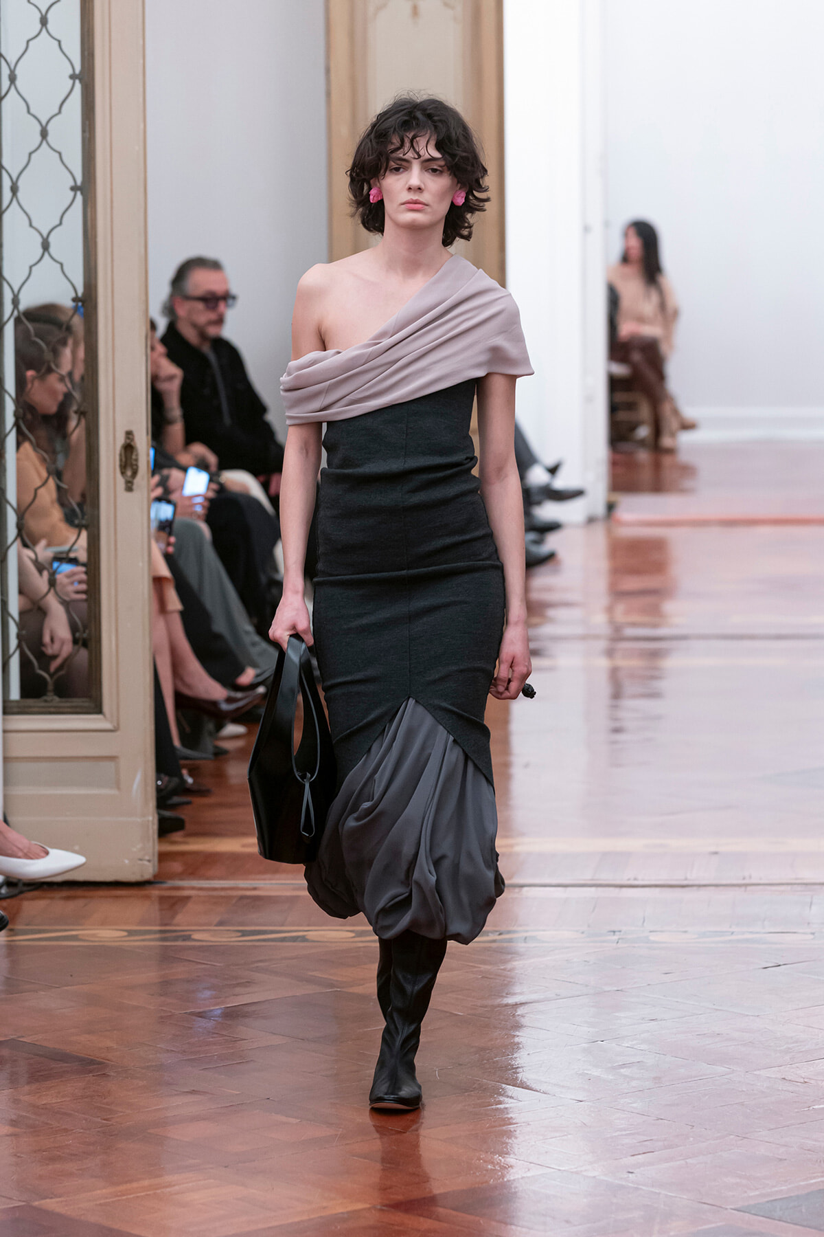 Model walks the runway in a dark, fitted dress with a pale pink off-shoulder drape and a sculpted gray bubble hem, carrying a black handbag.